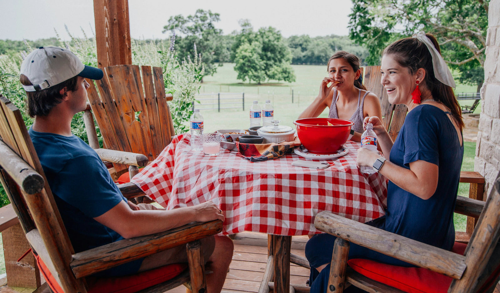 Trois personnes assises autour d'une table de pique-nique sur une terrasse en bois, partageant un repas en plein air avec une vue sur la campagne.